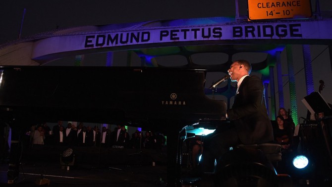 John Legend playing piano and singing in front of the Edmund Pettus Bridge